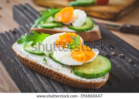 Tasty breakfast or snack toasts with cream cheese, fresh sliced cucumber and boiled egg, garnished with arugula leaves and spices on wooden cutting board. Selective focus