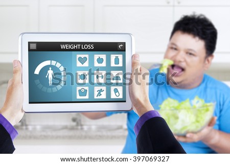 Picture of weight loss applications on the digital tablet screen and overweight person enjoying a bowl of salad in the kitchen