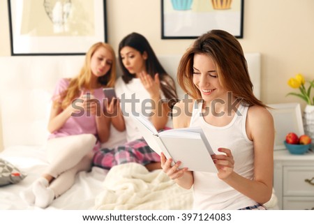 Two friends using mobile phones and one girl reading book on a bed in living room