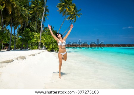 Happy young woman in white bikini runs on the tropical beach with her arms raised