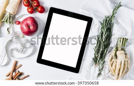 Tablet with a measuring tape, rosemary, nuts, parsley root, tomatoes, cinnamon and red apples on a white wooden background top view horizontal