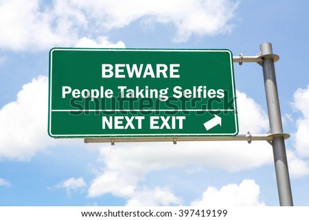 Green overhead road sign with a Beware of People Taking Selfies Next Exit concept against a partly cloudy sky background.