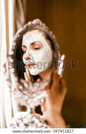 Woman applying mask of clay skin on face looking in mirror. Woman applying mask of clay skin on face looking in mirror.