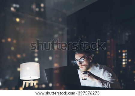 Bearded young businessman working on modern loft office at night. Man using contemporary notebook texting message, holding cup espresso, blurred background. Horizontal, film effect, bokeh