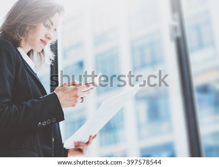 Photo business woman wearing suit, looking smartphone and holding documents in hands. Open space loft office. Panoramic windows background. Horizontal mockup. Film effect