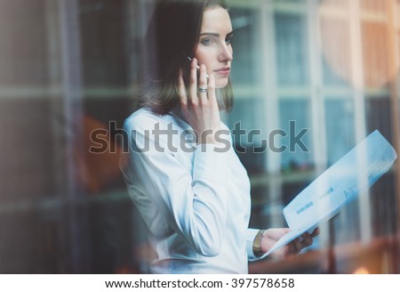 Photo business woman wearing white shirt, talking smartphone and holding documents in hands. Open space loft office. Panoramic windows background. Horizontal mockup, film effect