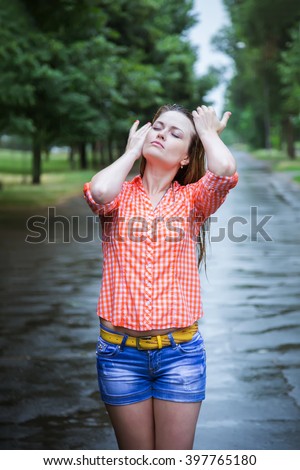 Young beautiful blond caucasian girl in shorts and shirt walking in the park in the summer warm rain barefoot through the puddles. Have fun and enjoy. Falling rain. Raindrops. Wet clothes