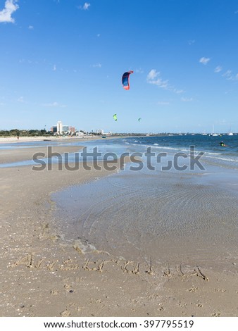 Australia inscription on the sand with shells on a large beautiful Australian beach, and unrecognizable people go kite surfing, and beautiful bright sails flying over blue sea, Australia