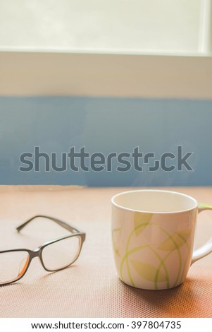 A cup of coffee placed  beside eyeglasses located by window in the morning.