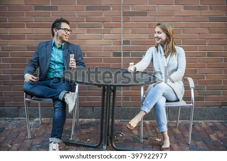 Two smiling young people with ice cream sitting at outdoor cafe table near brick wall having conversation