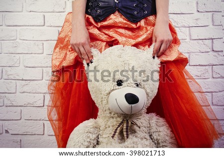 Girl in a lush orange skirt holding big soft teddy bear by the ears on white brick background