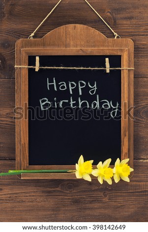 Happy Birthday written with chalk on aged blackboard with narcissus flowers hanging on wooden wall
