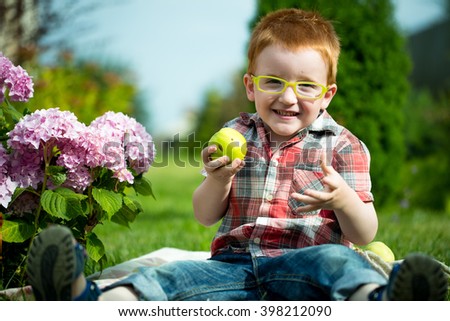 Happy laughing funny little red haired boy in stylish checkered shirt jeans and yellow glasses sitting outdoor on picnic with green apples near pink flowers on natural backdrop, horizontal picture