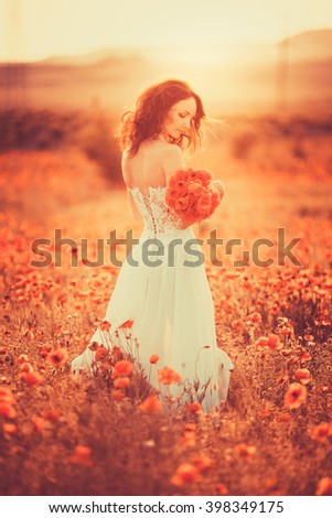 Beautiful dark-haired girl in a wedding dress of the bride at sunset on a summer evening in a field of poppies with a bouquet of red flowers, tenderness and femininity