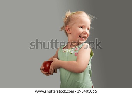 Happy Studio Portrait of Blond young girl wearing green dress and holding red apple at plain grey background 