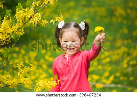 Portrait of funy little girl with yellow dandelions in hand. Beautiful smiling child playing with flowers on spring day in meadow. Baby holding bouquet of spring flowers at green grass background.