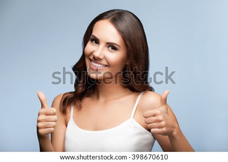 Portrait of happy smiling young beautiful woman in white casual clothing, showing thumbs up gesture, over grey background