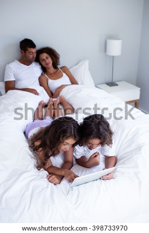 Brother and sister using digital tablet on bed while parents relaxing in background