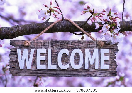 Wood welcome sign hanging from spring flowering tree branch; pink and white blossoms blurred in background