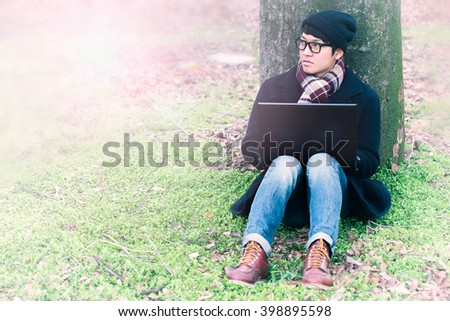 A nice looking  man is sitting against a tree in the grass, looking at his computer. He is relaxing, enjoying the shadow of the tree in a sunny day.