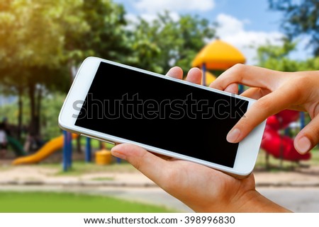 woman hand hold and touch screen smart phone, tablet,cellphone over blurred park and playground background.