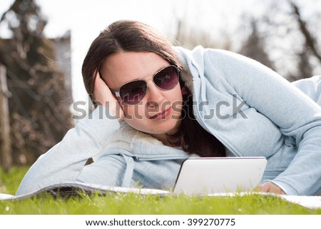 Pretty young girl / woman using white tablet with rose black sunglasses watching to film / serial and lying on grass during lovely summertime; looking happy; relax / rest time