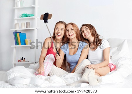 friendship, people, pajama party and technology concept - happy friends or teenage girls with smartphone and monopod taking selfie at home
