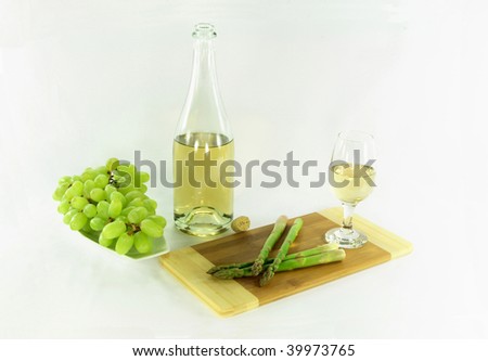 Picture of Still life - bottle and glass with white wine and sides grapes over the dish and asparagus over wooden cutting board over white background.
