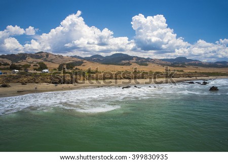 Ocean waves breaking against the shore under blue skies with white fluffy clouds.