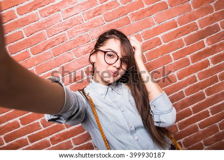 Beautiful young dark-haired girl in casual clothes and eyeglasses posing, smiling and making selfie, standing against brick wall