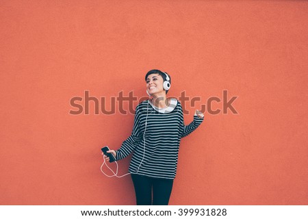 Knee figure of young beautiful caucasian woman listening music with headphones and smart phone hand hold, dancing, eyes closed smiling, leaning on a orange wall - music, happiness, technology concept Royalty-Free Stock Photo #399931828