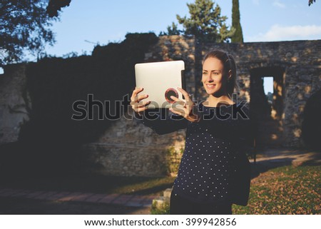 Cheerful hipster girl is making self portrait with touch pad during her walking in the fresh air in weekend. Smiling happy female student is photographing herself with front digital tablet camera