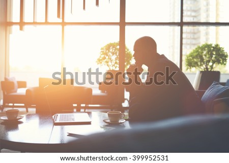 Silhouette of man managing director is thinking how to prevent the company's crisis,while is sitting with net-book in cafe. Male financier in watching world news on laptop computer during coffee break