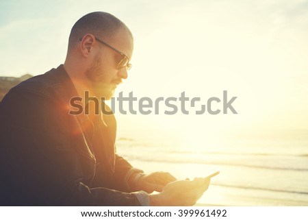 Young stylish hipster guy is searching information on web page on mobile phone,while relaxing during his spring weekend on the beach near sea with copy space background for your advertise text message