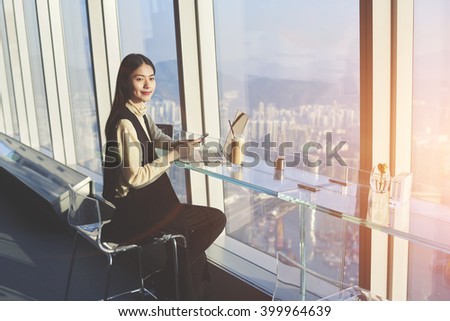 Young beautiful Asian female student with mobile phone in hands is looking at camera, while is sitting with laptop computer in co-working cafe with view of a business district outside the window