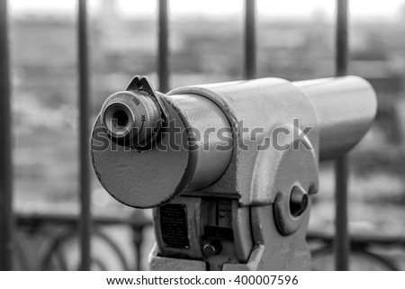 Use me now to see the World through me and dream...
The focus is on the viewfinder of a tower deck monocular with an unfocused city view and a fence in the background. In black and white.