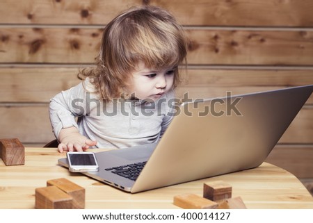 Cute funny little baby boy with long blonde curly hair playing on computer and mobile phone near toy building blocks indoor on wooden background, horizontal picture