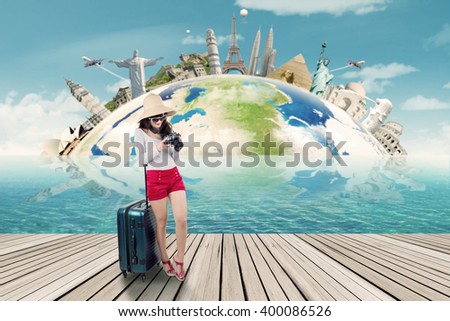 Beautiful woman holding a digital camera and standing on the jetty with luggage, shot with the world monuments background