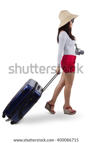 Picture of a female traveler walking in the studio while carrying a bag and a digital camera, isolated on white background