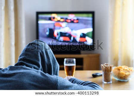 Tired hooded man falling asleep while watching TV (formula one race) in living room - stock photo
