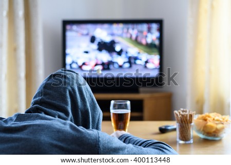 Tired hooded man falling asleep while watching TV (formula one race) in living room - stock photo