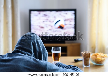 Hooded man watching TV (volleyball match) in living room with alcohol and snacks - stock photo