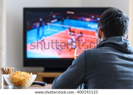 Tired man watching TV (volleyball match) in living room with alcohol and snacks - stock photo