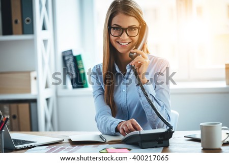I will connect you in one second! Cheerful young beautiful woman in glasses talking on the phone and looking at camera with smile while sitting at her working place Royalty-Free Stock Photo #400217065