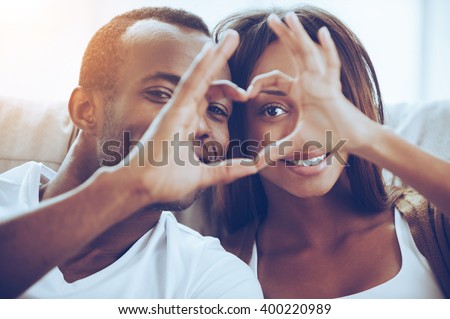 Love is in the air! Beautiful young African couple sitting close to each other and looking through a heart shape made with their fingers Royalty-Free Stock Photo #400220989