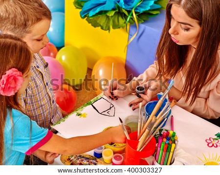 Children with teacher woman painting on paper at table  in  kindergarten . Balloons on floor.