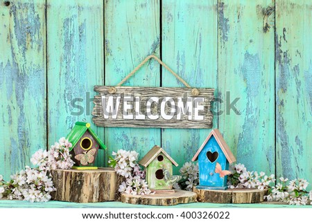 Welcome sign hanging over colorful birdhouses with butterfly on cedar logs by spring tree blossoms on antique rustic mint green wood background