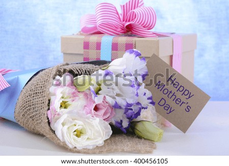 Beautiful Mother's Day lisianthus flowers wrapped in burlap and blue paper with gift box on white wood table and blue background, with gift tag.