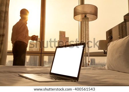 Laptop computer on white bed sheets in cozy room with copy space blank screen. Young businessman with cup of coffee standing at window looking at city scenery on the background. Motivation concept