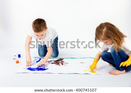 Two pretty child friends boy and girl in white shirts and blue jeans, trendy hair style, barefoot, drawing pictures on white sheet of paper by paints isolated on white. Studio shot.